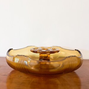 May include: A round amber-coloured glass flower frog bowl featuring a scalloped edge with vertical ribbed detailing. The centre of the bowl contains a raised, circular glass insert with multiple holes designed for arranging stems in a floral display.