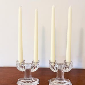 May include: Two clear glass double candle holders, each featuring a faceted starburst pattern on the cups and a vertical ribbed central column, standing on an octagonal base. Each holder contains two long, slender, off-white taper candles.
