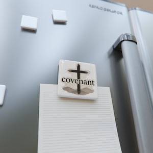May include: A square white refrigerator magnet featuring a black brushstroke cross graphic and the word covenant printed in black serif font. The magnet is positioned on a metallic surface, holding a piece of lined white paper in place.