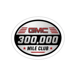 May include: An oval-shaped sticker with a brushed silver metallic background and a black border. The text GMC appears in red, followed by the number 300,000 in large silver font. Below this, the text MILE CLUB is flanked by two red stars.
