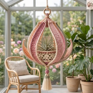 Könnte beinhalten: Ein hängender Pflanzenhalter aus Häkelarbeit mit einem von einer Lotusblume inspirierten Design, bestehend aus geschichteten Blütenblättern in Rosa, Creme und Salbeigrün. Er hängt an einem Holzring und endet in einer dekorativen Quaste.