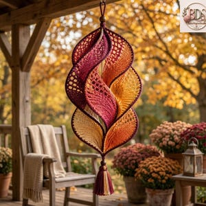 May include: A hanging spiral ornament crafted from crochet yarn in a gradient of deep burgundy, vibrant orange, and golden yellow. The structure features intricate openwork patterns and a twisted, leaf-like shape, finished with a decorative tassel.