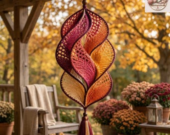 Peut inclure: Une décoration suspendue en spirale réalisée en fil crocheté avec un dégradé de bordeaux profond, d'orange vif et de jaune doré. La structure présente des motifs ajourés complexes et une forme torsadée en feuille, terminée par un pompon.