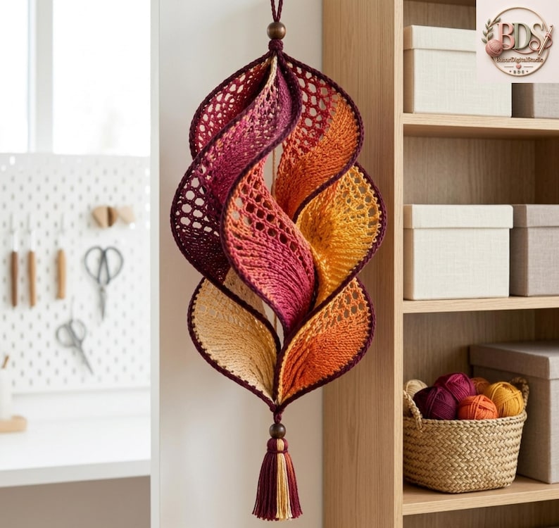 K&ouml;nnte beinhalten: Eine h&auml;ngende H&auml;keldekoration mit einem Spiraldesign und einem Farbverlauf von Dunkelviolett und Magenta bis hin zu leuchtendem Orange und Gelb. Das St&uuml;ck ist oben mit einer dunkelbraunen Holzperle und unten mit einer passenden Quaste versehen.