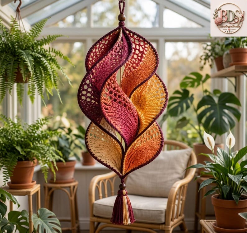 K&ouml;nnte beinhalten: Ein h&auml;ngendes H&auml;kel-Spiralornament mit einem Farbverlauf von tiefem Burgunderrot zu leuchtendem Orange und Gelb. Das St&uuml;ck weist eine durchbrochene Netzstruktur, eine zentrale Holzperle an der Oberseite und eine mehrfarbige Quaste am unteren Ende auf.