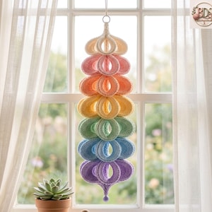 May include: A hanging crochet decoration featuring a vertical stack of six rounded, hollow, teardrop-shaped segments in a gradient of colors including white, pink, orange, yellow, green, blue, and purple, suspended by a thin white string.