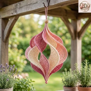 May include: A hanging crochet ornament featuring a spiraling, three-dimensional shape. The piece is crafted with yarn in gradient shades of cream, soft pink, and deep rose, suspended from a single loop with a small wooden bead at the top and bottom.