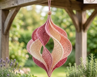 Könnte beinhalten: Ein hängendes Häkelornament in einer spiralförmigen, dreidimensionalen Form. Das Stück ist aus Garn in abgestuften Farbtönen von Creme, zartem Rosa und tiefem Rosé gefertigt und an einer Schlaufe mit einer kleinen Holzperle oben und unten befestigt.