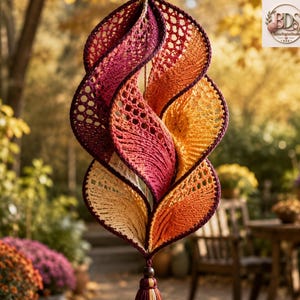 K&ouml;nnte beinhalten: Ein h&auml;ngendes Spiralornament aus geh&auml;keltem Garn in einem herbstlichen Farbverlauf, der von tiefem Burgunderrot und Dunkelviolett oben zu leuchtendem Orange und sanftem Gelb unten &uuml;bergeht, abgeschlossen mit einer kleinen dunklen Quaste.