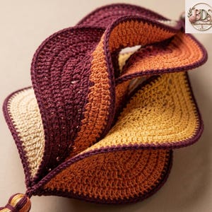 K&ouml;nnte beinhalten: Ein dreidimensionales H&auml;kelobjekt mit einem gedrehten Spiraldesign. Das St&uuml;ck besteht aus Garn in den Farben Dunkelburgunder, gebranntem Orange und Creme, wobei jeder Abschnitt mit einem dunkelburgunderfarbenen Rand und einer kleinen Quaste versehen ist.