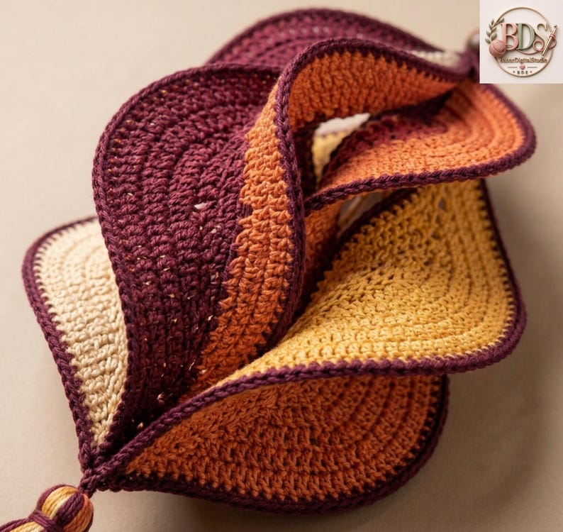 K&ouml;nnte beinhalten: Ein dreidimensionales H&auml;kelobjekt mit einem gedrehten Spiraldesign. Das St&uuml;ck besteht aus Garn in den Farben Dunkelburgunder, gebranntem Orange und Creme, wobei jeder Abschnitt mit einem dunkelburgunderfarbenen Rand und einer kleinen Quaste versehen ist.
