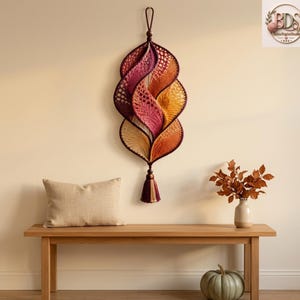 K&ouml;nnte beinhalten: Ein vertikaler Wandbehang bestehend aus einer Reihe miteinander verbundener, blattf&ouml;rmiger H&auml;kelsegmente in einem Farbverlauf von Dunkelviolett, Magenta, Orange und Creme. Das St&uuml;ck ist an einer Schlaufe aufgeh&auml;ngt und mit einer Quaste versehen.
