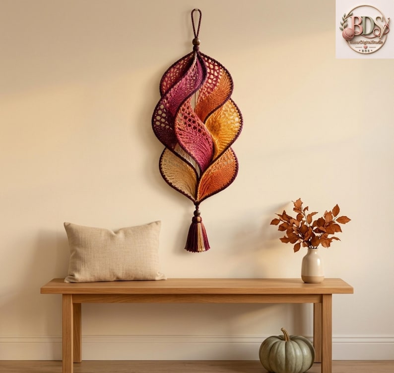 K&ouml;nnte beinhalten: Ein vertikaler Wandbehang bestehend aus einer Reihe miteinander verbundener, blattf&ouml;rmiger H&auml;kelsegmente in einem Farbverlauf von Dunkelviolett, Magenta, Orange und Creme. Das St&uuml;ck ist an einer Schlaufe aufgeh&auml;ngt und mit einer Quaste versehen.