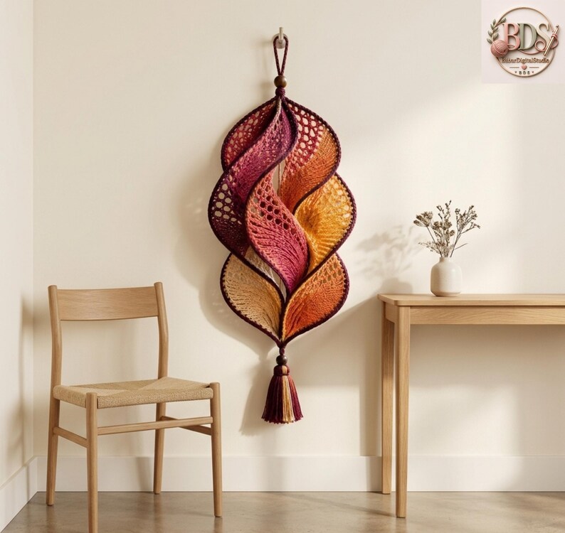 K&ouml;nnte beinhalten: Ein vertikaler Wandbehang mit einem Spiralmuster aus geh&auml;keltem Garn in einem Farbverlauf von Dunkelviolett, Magenta, Orange und Creme. Das St&uuml;ck ist an einer Holzperle aufgeh&auml;ngt und mit einer passenden, mehrfarbigen Garnquaste versehen.