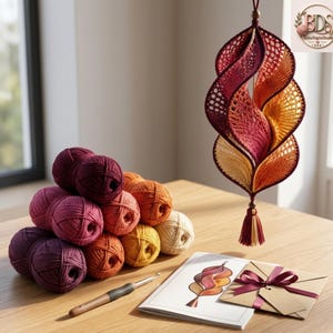 K&ouml;nnte beinhalten: Ein h&auml;ngendes Spiralornament aus geh&auml;keltem Garn in Farbverl&auml;ufen von Dunkelviolett, Rot, Orange und Gelb. Daneben liegen ein Stapel passender Garnkn&auml;uel, eine Metallh&auml;kelnadel, ein gedrucktes Anleitungsheft und ein Geschenkumschlag mit einer Schleife.