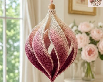 Könnte beinhalten: Ein hängendes Häkelornament mit spiralförmigem, tropfenförmigem Design. Das Garn verläuft farblich von einem hellen Creme oben bis zu einem tiefen Burgunderrot unten. Eine kleine Holzperle sitzt oben und eine weiße Perle hängt an der Unterseite.