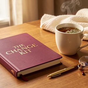 Könnte beinhalten: Ein kastanienbraunes Hardcover-Buch mit dem Titel THE CHANGE KIT in goldener Schrift, begleitet von einer weißen Keramiktasse mit einem dampfenden Getränk, einem goldfarbenen Kugelschreiber und einer Brille mit rundem Rahmen und dünnen Bügeln.