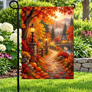 May include: A rectangular garden flag featuring a vibrant autumn landscape with a stone path, two glowing black street lamps, orange pumpkins, and a small house near a bridge, all surrounded by trees with red, orange, and yellow fall foliage.