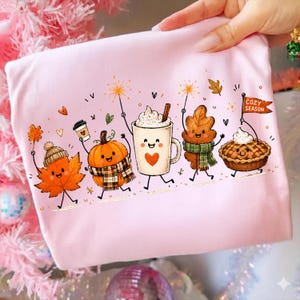 May include: A light pink t-shirt featuring a colorful graphic of animated autumn characters including a maple leaf in a beanie, a pumpkin with a plaid scarf, a mug of hot cocoa with a heart, an oak leaf with a scarf, and a pie with a flag reading COZY SEASON.