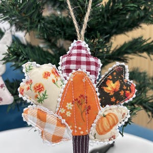 May include: A fabric maple leaf ornament featuring a patchwork design of various autumn-themed patterns including red gingham, orange floral, and pumpkin prints. The leaf is edged with white lace trim and includes a jute twine loop for hanging.