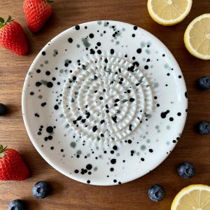 Può includere: Un piatto grattugia rotondo in ceramica bianca con un motivo a macchie nere e grigie. Il centro del piatto presenta un design circolare in rilievo e testurizzato per grattugiare il cibo, circondato da un bordo liscio. Il piatto è circondato da frutta.