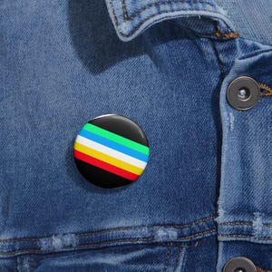Può includere: Una spilla circolare con un motivo a strisce orizzontali nei colori nero, verde, azzurro, bianco, giallo e rosso. La spilla è fissata su un tessuto denim blu con un bottone in metallo visibile e cuciture in evidenza.