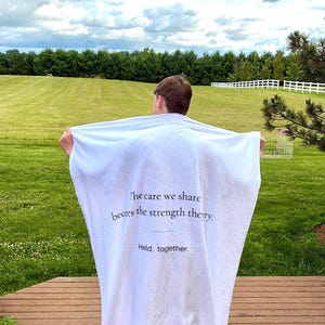 May include: A white rectangular blanket featuring black printed text that reads: The care we share becomes the strength they. Held, together. The fabric has a soft, plush texture and is held open to display the full message printed in the center.