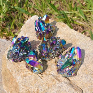May include: Five clusters of iridescent titanium aura quartz crystals featuring a metallic rainbow finish with vibrant shades of blue, purple, green, and gold. Each cluster displays sharp, pointed crystalline structures with a reflective surface.
