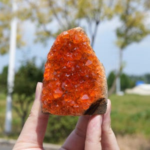 Könnte beinhalten: Ein rohes Mineralexemplar mit einer Ansammlung von leuchtend orangefarbenen, durchscheinenden Kristallen mit scharfen, kantigen Facetten. Die Kristallformation ist auf einer natürlichen, dunkelbraunen, felsigen Basis befestigt und zeigt einen deutlichen Kontrast zwischen der hellen, reflektierenden Kristalloberfläche und der rauen, erdigen Textur des Steinfundaments.
