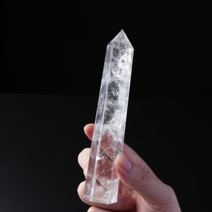 May include: A clear, hexagonal quartz crystal point with a pointed top and a faceted, translucent body. The crystal displays natural internal inclusions and fractures throughout its structure, reflecting light against a dark, solid background.