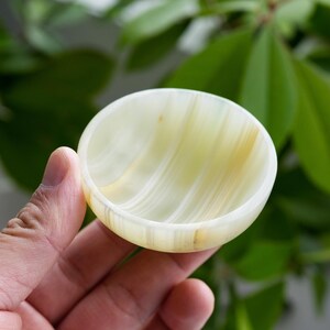 Puede incluir: Un cuenco pequeño, redondo y poco profundo tallado en piedra pulida con bandas de color verde claro y crema. El cuenco presenta capas lisas y translúcidas con sutiles estrías naturales fluidas en todo el material, creando un aspecto suave y veteado.
