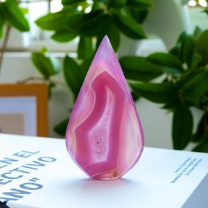 May include: A teardrop-shaped polished agate stone featuring vibrant shades of pink and translucent white banding. The centre of the stone displays a textured, crystalline geode cavity, showcasing natural mineral formations within the smooth surface.