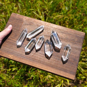 Pode incluir: Seis pontas de cristal de quartzo transparente de terminação dupla dispostas numa tábua de madeira retangular castanho-escura. Cada cristal apresenta uma forma hexagonal com extremidades pontiagudas, transparência natural e facetas.