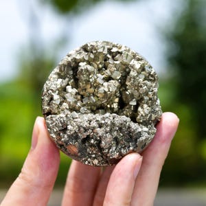 Puede incluir: Un racimo de pirita esférico que presenta una disposición densa de cristales cúbicos metálicos de color latón. El espécimen mineral muestra una superficie texturizada y multifacética con un brillo dorado reflectante en toda la formación redondeada.
