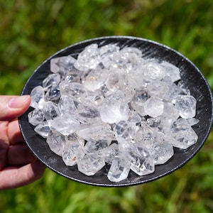 Op de afbeelding: Een verzameling kleine, heldere, ruwe kwartskristallen opgestapeld op een rond, zwart, getextureerd bord. De kristallen zijn doorschijnend, veelzijdig en variëren in vorm en grootte, waarbij ze het licht reflecteren op hun grillige, natuurlijke oppervlakken.