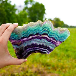May include: A large, flat slice of fluorite crystal featuring distinct, jagged bands of deep purple, vibrant teal, and translucent white. The mineral specimen displays a natural, irregular geometric shape with sharp, angular edges and a smooth surface.