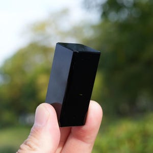 Può includere: Un solido blocco rettangolare in pietra ossidiana nera lucidata. L'oggetto presenta una superficie liscia e riflettente con bordi netti e puliti, e un colore nero opaco e profondo in tutta la sua struttura, mostrando un aspetto uniforme e denso.