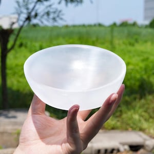 Puede incluir: Un cuenco de selenita blanca translúcida con un borde redondeado suave y un acabado pulido. El cuenco tiene una forma circular y una apariencia sólida y opaca que permite que la luz pase a través de sus bordes, creando un brillo suave en la superficie.