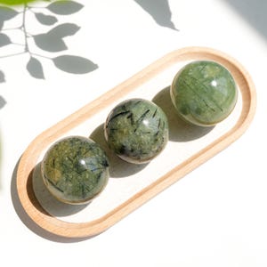 May include: Three polished spherical stones with a translucent sage green base and distinct black needle-like inclusions. The spheres are arranged in a line on an oval-shaped wooden tray with a light-coloured fabric insert base.