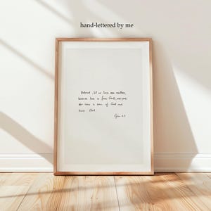May include: A framed wall art print featuring a light beige background with black hand-lettered text that reads: Beloved, let us love one another, because love is from God; everyone who loves is born of God and knows God. 1 John 4:7.