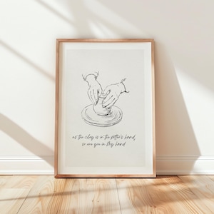 May include: A framed wall art print featuring a black line drawing of hands shaping a clay pot on a pottery wheel. Below the illustration, the text reads: as the clay is in the potter's hand, so are you in My hand. The print has a light background.