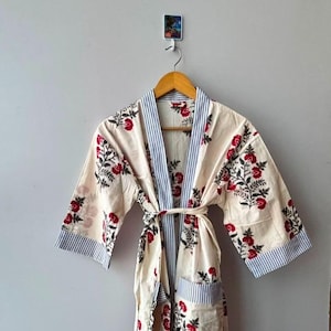 May include: A cream-coloured robe featuring a red floral print with green leaves. The garment includes blue and white striped trim along the front opening, sleeve cuffs, and two front pockets. A matching fabric tie belt is secured at the waist.
