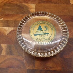 May include: A round clear glass coaster with a scalloped edge featuring a central gold-toned circular emblem. The emblem displays a graphic of a sailboat and sun with the text: Honoring Senator Claiborne Pell, Rhode Island, April 26, 1998, USAD.