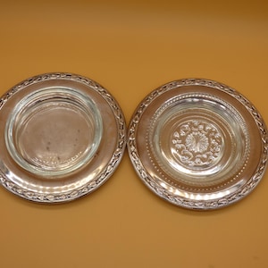 May include: Two round coasters featuring a clear glass center insert surrounded by a metallic silver-toned rim. The left coaster has a smooth glass surface, while the right coaster displays a decorative embossed floral pattern in the glass center.