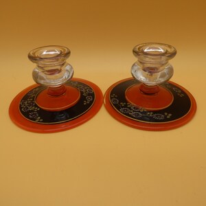 May include: A pair of glass candle holders featuring clear glass tops and wide, circular bases. The bases are orange with a black inner ring decorated with a delicate white floral pattern and thin gold-colored circular trim accents.