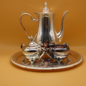 May include: A silver-plated tea service set consisting of a tall teapot with a lid, a lidded sugar bowl, and an open creamer, all arranged on a round silver-plated serving tray. Each piece features ornate curved handles and decorative feet.