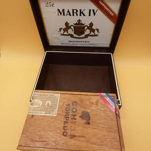 May include: An open dark brown box with a white interior lid featuring the text MARK IV, 25c, and a crest with two horses. In front sits a closed wooden box labeled COHIBA TORPEDO with a rectangular stamp and a red, white, and blue seal.