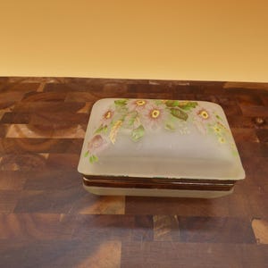 May include: A rectangular frosted glass trinket box featuring a painted floral design with pink and yellow flowers and green leaves on the lid. The box has a metallic band around the center where the lid meets the base, creating a hinged closure.
