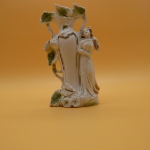 May include: A white porcelain vase featuring a sculpted figure of a person standing beside the vessel. The vase is adorned with raised green leaf motifs and small white flowers at the base, accented with subtle gold trim along the edges.