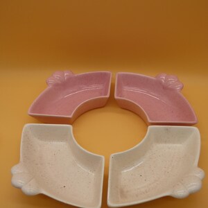 May include: A set of four fan-shaped ceramic dishes arranged in a circular pattern. Two dishes are light pink with a speckled texture, and two dishes are off-white with a speckled texture. Each dish features a decorative shell-like handle detail.